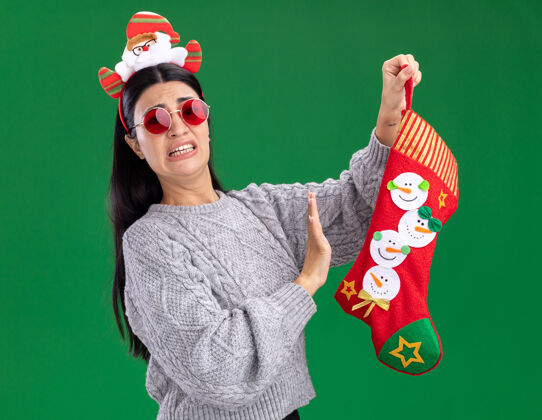 手势恼怒的年轻白人女孩戴着圣诞老人的头带戴着眼镜拿着圣诞长袜看着相机做着拒绝的手势隔离在绿色的背景上眼镜圣诞快乐圣诞老人