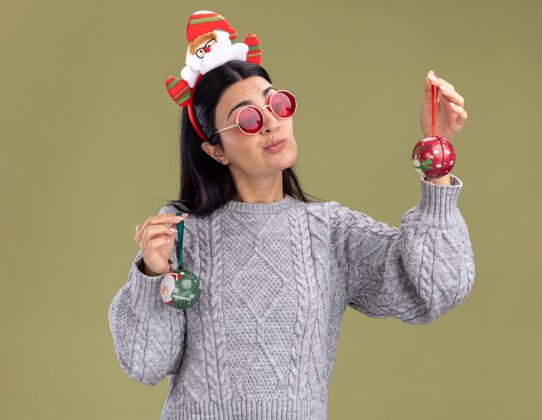 圣诞饰品可疑的年轻白人女孩戴着圣诞老人的头带 戴着眼镜 手里拿着圣诞饰品 看着其中一个孤立的橄榄绿背景圣诞眼镜抱着
