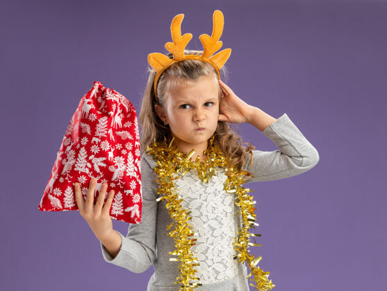 包皱眉的小女孩戴着圣诞发箍 脖子上戴着花环 手放在头上 手放在蓝色背景上花环蓝色箍