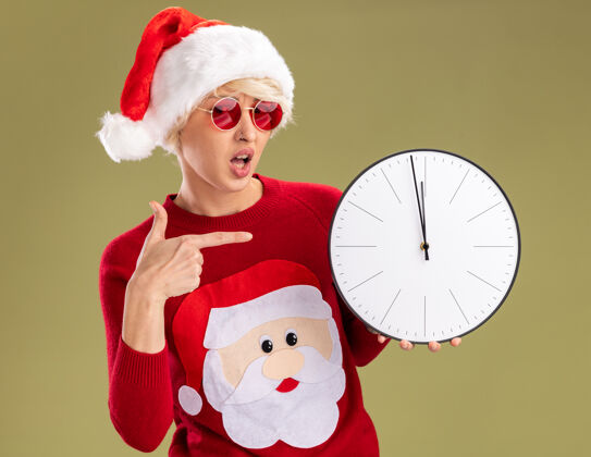 女人令人印象深刻的年轻金发女人戴着圣诞帽和圣诞老人的圣诞毛衣 戴着眼镜 指着时钟 看着橄榄绿背景上孤立的摄像机帽子时钟毛衣