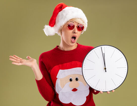 年轻令人印象深刻的年轻金发女人戴着圣诞帽和圣诞老人的圣诞毛衣戴着眼镜拿着时钟看着相机显示空手孤立在橄榄绿的背景圣诞手眼镜