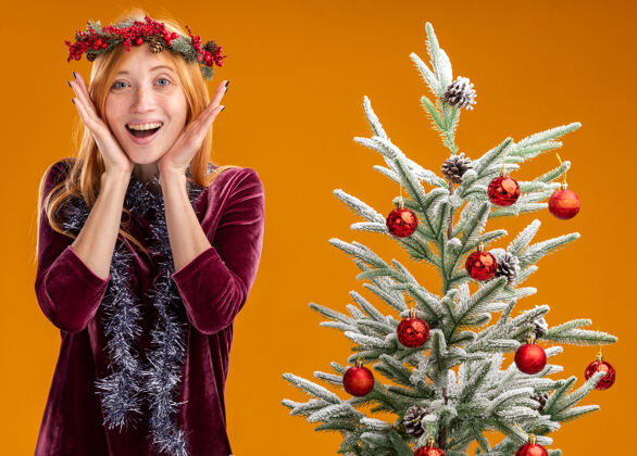 手兴奋的年轻漂亮女孩站在圣诞树旁 身穿红色连衣裙 脖子上戴着花环 手牵着手 脸孤立在橙色背景上圣诞快乐年轻红色