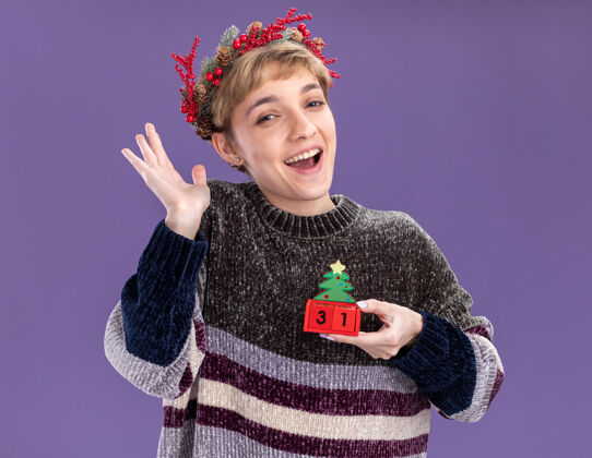 年轻快乐的年轻漂亮女孩戴着圣诞花环手持圣诞树玩具和日期看着相机显示空手孤立在紫色背景上欢乐空举行
