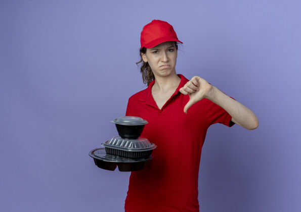 漂亮年轻漂亮的送货女孩 穿着红色制服 戴着帽子 拿着食物容器 拇指朝下 紫色背景上有复印空间容器向下按住