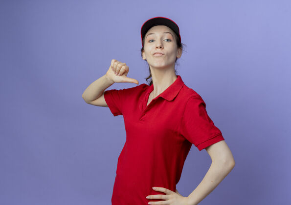 信心自信的年轻漂亮的送货女孩 穿着红色制服 戴着帽子 手放在腰上 指着自己孤立的紫色背景和复制空间漂亮年轻手