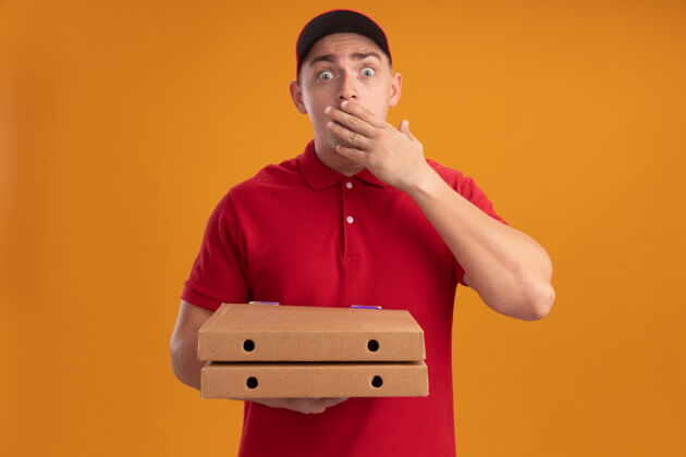递送被吓到的年轻送货员穿着制服 戴着帽子 拿着披萨盒 嘴上蒙着一只手 隔离在橙色的墙上表情嘴感觉