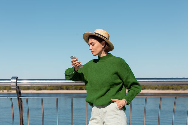 聊天穿着绿色休闲毛衣 戴着帽子的时尚女士在户外的桥上 在温暖阳光明媚的夏日里欣赏河景 手持手机微笑电话蓝色休闲
