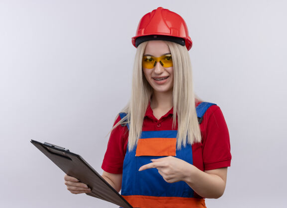 建筑商令人印象深刻的年轻金发工程师建设者女孩制服戴着牙套戴着安全眼镜拿着剪贴板 指着它在孤立的白色空间制服穿年轻