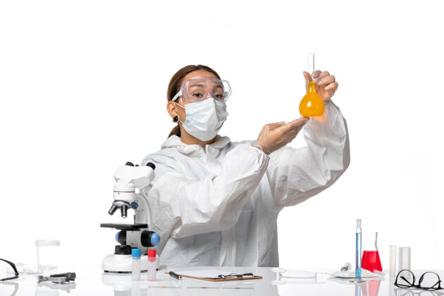 瓶子正面图：女医生 穿着特制的工作服 戴着口罩 拿着一个装有黄色溶液的瓶子 背景是白色的冠状病毒-大流行性冠状病毒药病毒前面