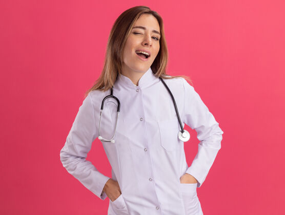 手眨眼的年轻女医生穿着医用长袍 听诊器把手放在口袋里 隔离在粉红色的墙上听诊器长袍人