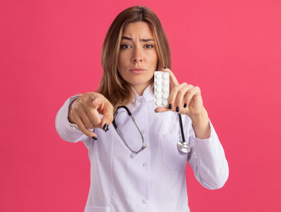 表情自信的年轻女医生穿着带听诊器的医用长袍 手持药片 站在隔离在粉红色墙上的前面女性长袍年轻人