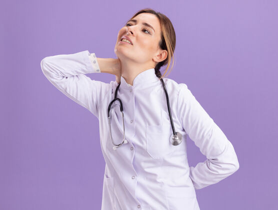 感情受伤的年轻女医生穿着医用长袍 手持听诊器 抓住了隔离在紫色墙上的疼痛的脖子人抓取疼痛