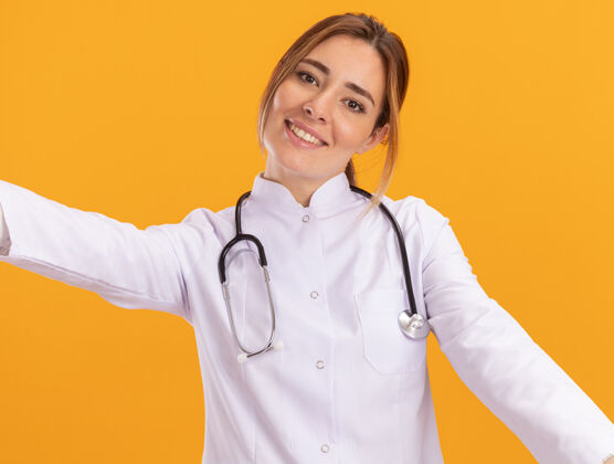 衣服面带微笑的年轻女医生穿着医用长袍 听诊器举着隔离在黄色墙壁上的前部医学感觉脸