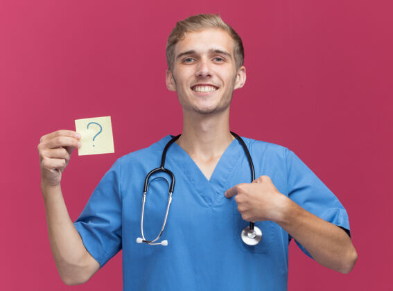 公民微笑的年轻男医生穿着医生制服手持听诊器问号指着隔离在粉红色墙上的自己衣服微笑年轻人