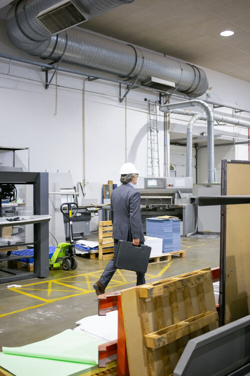 安全穿着正装戴着安全帽的成熟工程师走在工厂的地板上 带着工具的手提箱劳工头盔成熟