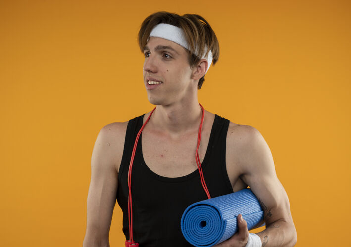 运动型很高兴年轻的运动型家伙看着一边 脖子上系着跳绳 手里拿着隔离在橙色墙上的瑜伽垫绳子请壁板