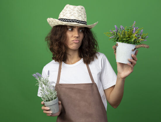 花园一个穿着制服的年轻女园丁戴着园艺帽 手拿花盆 看着隔离在绿色上的花朵不愉快穿花