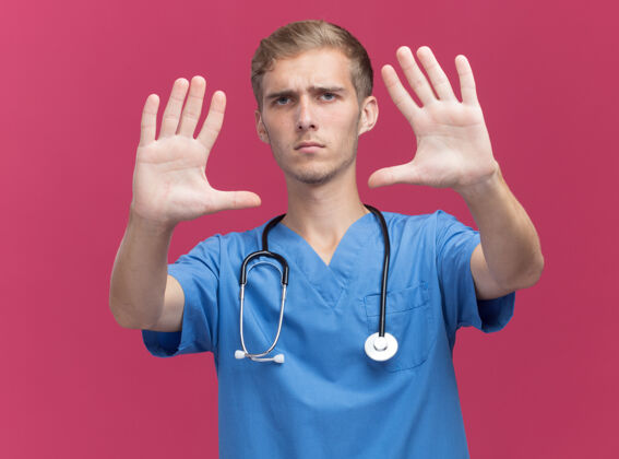表演严格的年轻男性医生穿着医生制服 听诊器显示停止手势隔离在粉红色的墙上听诊器男性制服