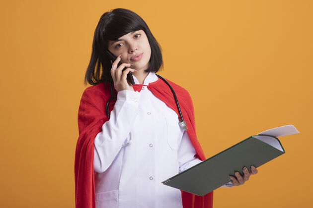 年轻年轻的超级英雄女孩戴着听诊器 穿着医用长袍和披风 拿着剪贴板 在橙色的墙上讲电话说话长袍电话