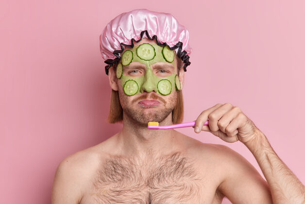 美女不高兴的男人用黄瓜片在脸上涂上绿色滋养面膜 让皮肤焕发青春伤心地看着相机刷牙站在粉色背景下 赤裸上身健康粉色健康