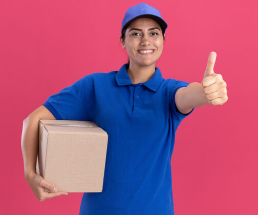 帽子微笑着的年轻送货女孩穿着制服 戴着帽子的盒子 在粉红色的墙上孤立地竖起大拇指制服显示微笑