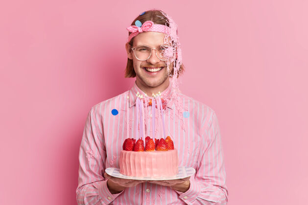 乐趣摄影棚拍摄的“快乐男人”庆祝生日手持美味的草莓蛋糕与穿着节日服装的客人见面 粉色背景下是孤立的庆祝脸高兴