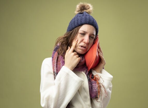 穿哭泣的生病的年轻女孩穿着白色长袍 戴着冬天的帽子 戴着围巾 把热水袋放在脸颊上 手指放在脸颊上 隔离在橄榄绿上疾病脸颊绿