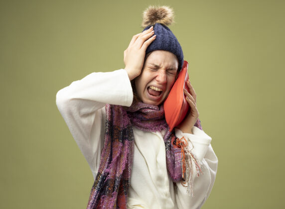 头病痛的年轻女孩穿着白色长袍 戴着冬天的帽子 戴着围巾 把热水袋放在脸颊上 把头孤立在橄榄绿上水痛帽子