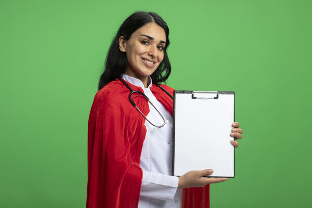 听诊器微笑着的年轻超级英雄女孩穿着医用长袍 听诊器拿着剪贴板隔离在绿色剪贴板年轻超级英雄