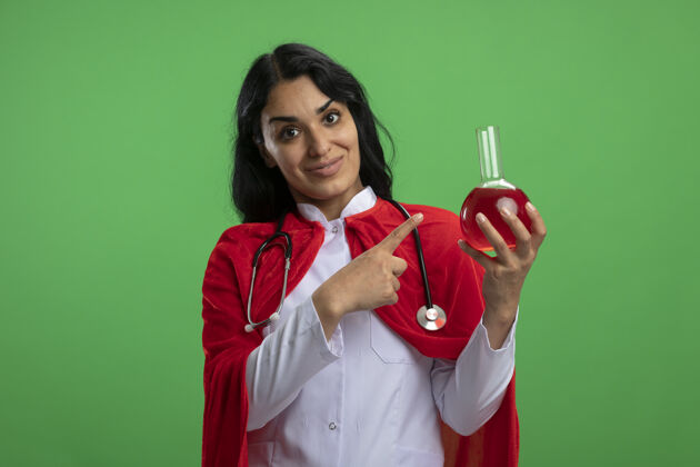 填补高兴的年轻超级英雄女孩穿着医用长袍 手持听诊器 指着装满红色液体的化学玻璃瓶 绿色隔离化学医疗瓶子