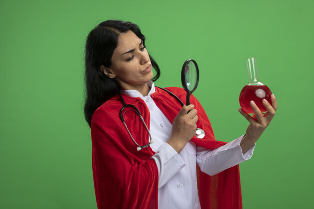 液体沉思的年轻超级英雄女孩穿着医用长袍 手持听诊器 看着装满红色液体的化学玻璃瓶 放大镜隔离在绿色上思考放大镜长袍