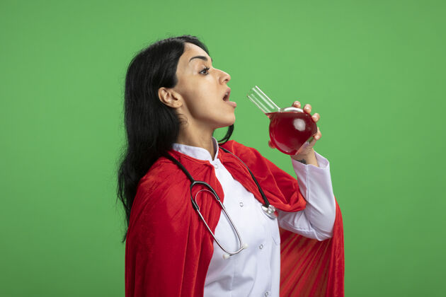 红色站在侧视图年轻的超级英雄女孩穿着医用长袍与听诊器举行和饮料化学玻璃瓶装满红色液体隔离在绿色持有听诊器瓶子