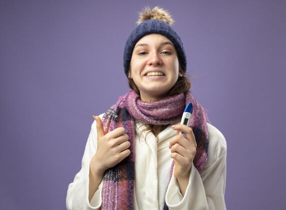 抱着带着微笑的生病的年轻女孩 穿着白色长袍 戴着冬天的帽子 戴着围巾 拿着温度计 大拇指朝上 孤立地站在紫色地上年轻长袍拇指