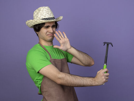 花园年轻的男园丁穿着制服 戴着园艺帽 拿着锄头看耙子 孤立地站在紫色地上紫色男性持有