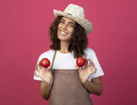 穿微笑的年轻女园丁穿着制服 戴着园艺帽 手里拿着粉红色的西红柿园艺制服番茄