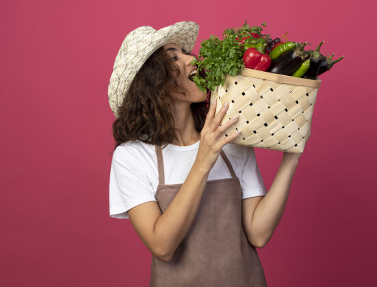年轻的高兴的年轻女园丁穿着制服 戴着园艺帽 捧着菜篮子看着肩上孤立的粉红色篮子拿着蔬菜
