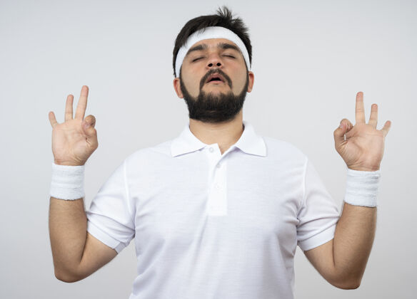 手势年轻的运动型男子 戴着头带和腕带 闭着眼睛 在白色的墙上显示出冥想的姿势封闭男人年轻