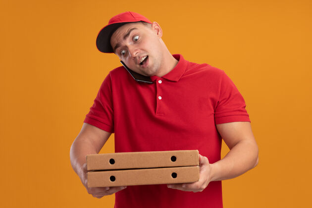 送货惊讶的年轻送货员穿着制服 戴着帽子 拿着披萨盒 隔着橙色的墙上讲电话盒子电话惊喜