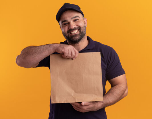 送货微笑的中年送货员穿着制服 戴着帽子 手里拿着纸食品包 隔离在黄色的墙上拿着制服微笑