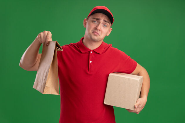 悲伤悲伤的年轻送货员穿着制服 戴着帽子 手里拿着纸食品包 盒子被隔离在绿色的墙上男人年轻包装