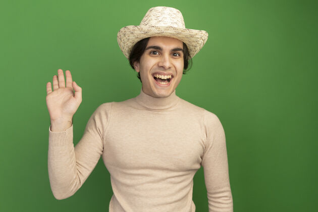 穿带着微笑的年轻帅哥戴着帽子在绿色的墙上显示问候的手势帅气年轻手势