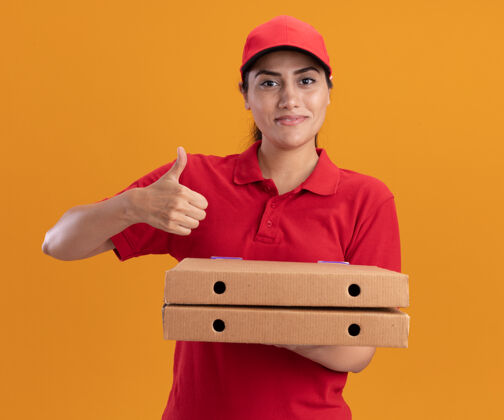 盒子微笑的年轻送货女孩穿着制服 戴着帽子 拿着披萨盒 在橙色的墙上孤立地竖起大拇指微笑年轻送货