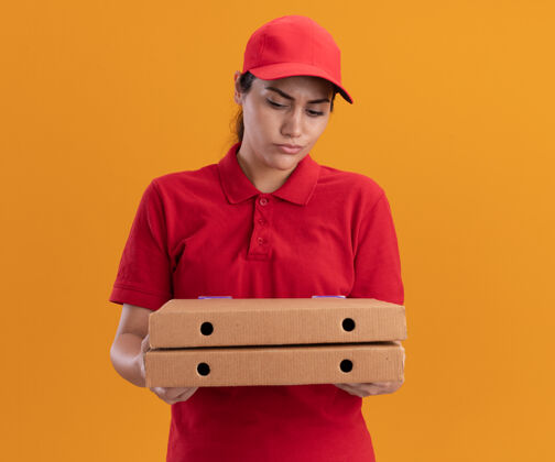 盒子困惑的年轻送货女孩穿着制服 戴着帽子 看着隔离在橙色墙上的比萨饼盒披萨送货女孩