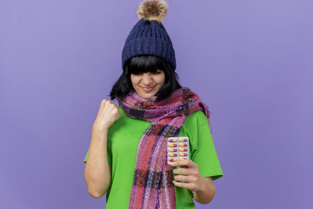 手势高兴的年轻生病的女人戴着冬天的帽子和围巾拿着一包胶囊做是的手势隔离在紫色的墙上复制空间人穿公民