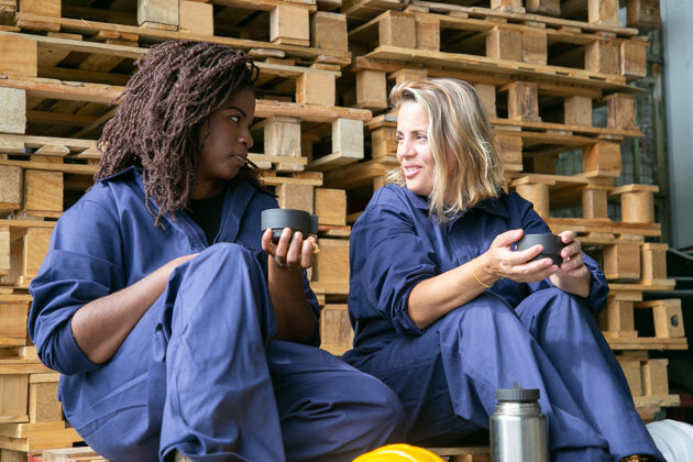 坐着积极的女工一边聊天一边喝咖啡 坐在仓库的木平台上生产木头职业