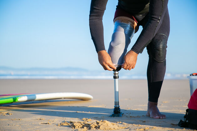 运动冲浪者穿着潜水服 站在沙滩上的冲浪板旁 调整绑在腿上的假肢受伤腿站立