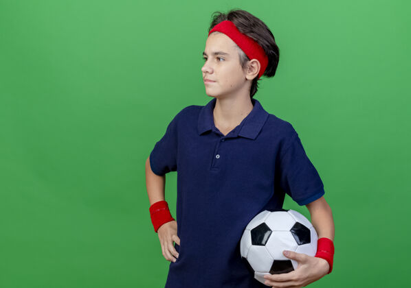 穿自信的年轻帅气的运动男孩戴着头带和护腕 戴着牙套 手里拿着足球 手放在腰上 看起来笔直 隔离在绿色的墙上 有复制空间市民表情感觉