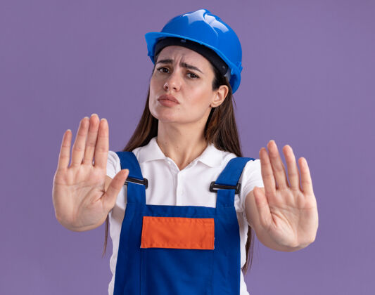 严格穿制服的严格的年轻建设者妇女在紫色的墙上显示停止手势表演停下女人