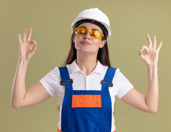 关闭喜欢闭上眼睛的年轻建筑女 穿着制服 戴着眼镜 在橄榄绿的墙上显示出冥想的姿势建筑工人请女士