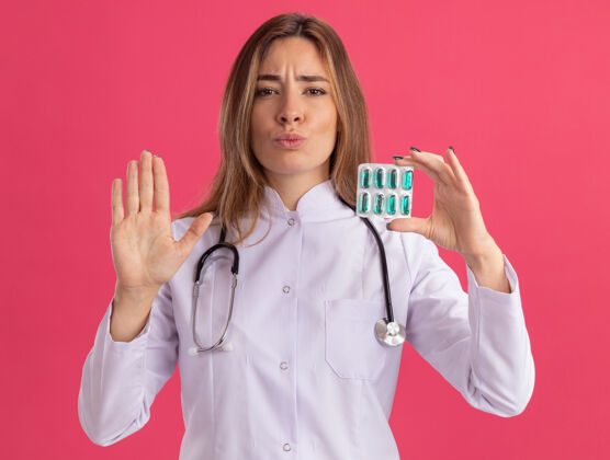长袍年轻严格的女医生 穿着医用长袍 手持听诊器 在粉色墙上显示停止手势医生听诊器年轻